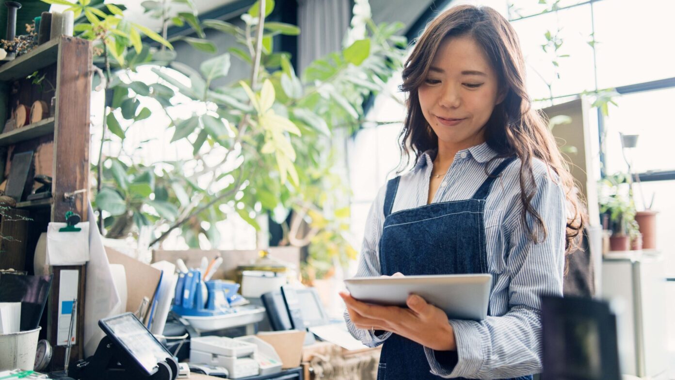 A woman works on her small business. She's wearing an apron and smiling while using an iPad to explore financing solutions.