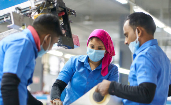 A group of workers collaborate in a factory wearing smart blue shirts and face masks.
