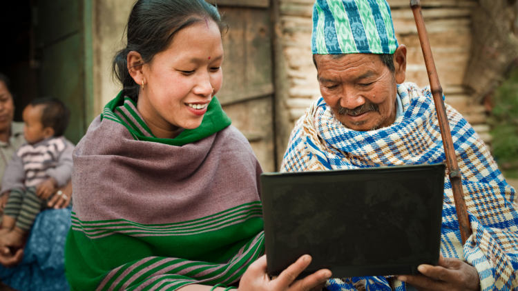 A woman helps a man use a laptop. They're in a rural village and both wearing traditional dress.