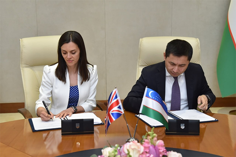 A man and a woman, both wearing smart business dress, sign papers at a table. The table has a UK flag and Uzbekistan flag.