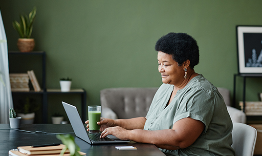 A woman works from home on a laptop. She's smiling and drinking a green juice.