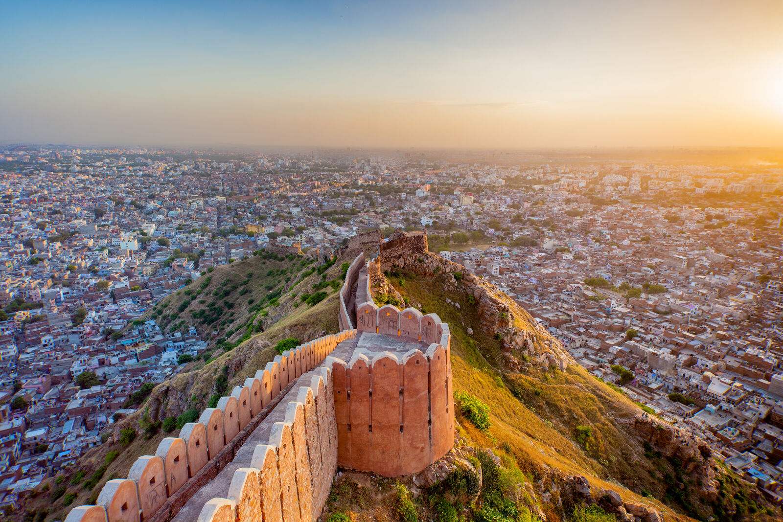 A view of historic fort wall winding along a hillside overlooking a densely populated city at sunset. 