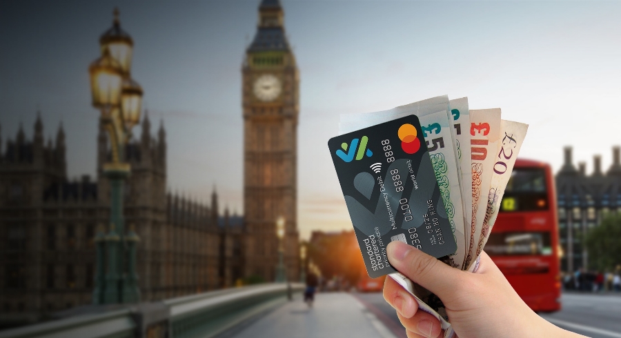 A hand holding a Multi-Currency Mastercard Debit Card and foreign currency against London scenery