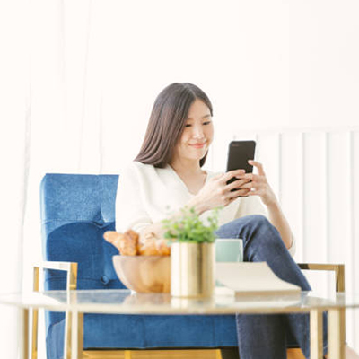A lady with long hair, dressed in a white sweater and blue jeans, sits on a blue sofa and holding a mobile phone