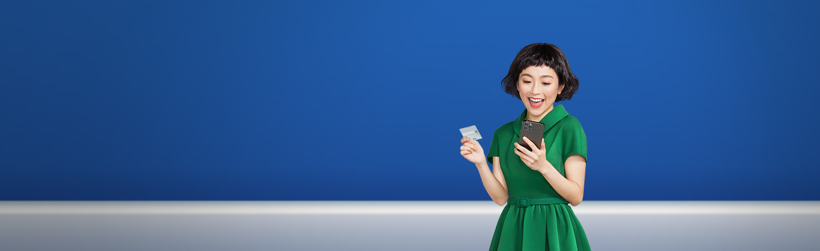 一名身穿綠色連身裙的年輕女士開心地使用她的手機及拿着渣打Smart卡