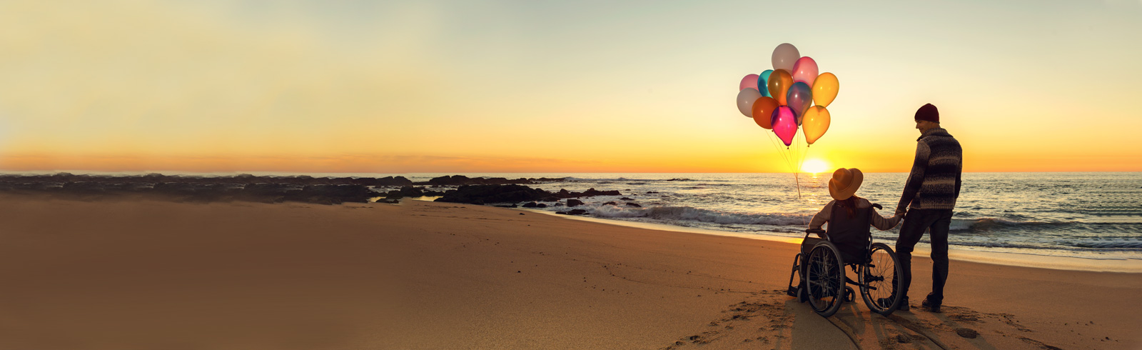 一位女士坐著輪椅並拿著氣球,與伴侶在海灘上觀賞日落