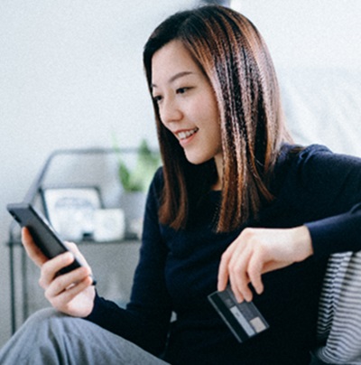 Beautiful smiling young asian woman sitting on the floor in the bedroom shopping online with smartphone and making mobile payment with credit card on hand at cozy home technology makes life so much easier